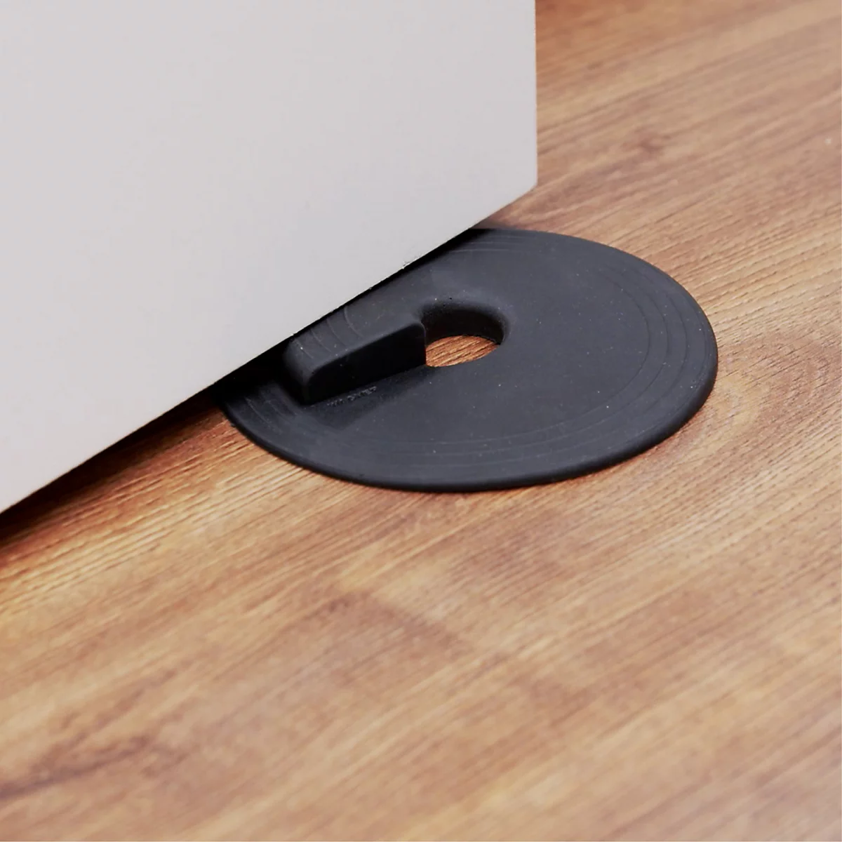 Un butoir de porte en caoutchouc noir devant une porte blanche sur un sol en bois. Le butoir de porte a une forme ronde avec un trou et une saillie.