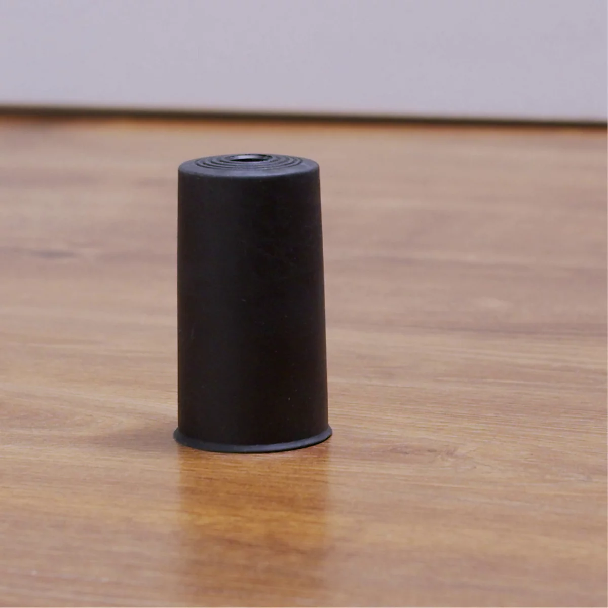 Un patin de meuble noir et cylindrique se dresse sur un sol en bois, qui a une ligne noire d'un côté.