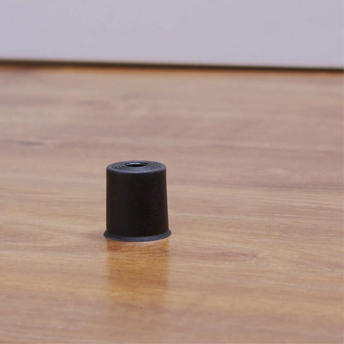 Couvercle cylindrique noir pour un pied de meuble, sur un sol en bois clair. L'objet est placé au centre.