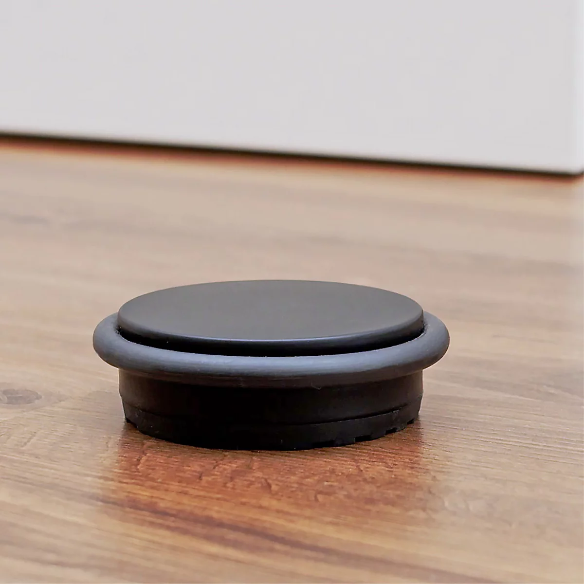 Un butoir de porte noir sur un plancher en bois. Le butoir a une forme ronde et un anneau en caoutchouc.