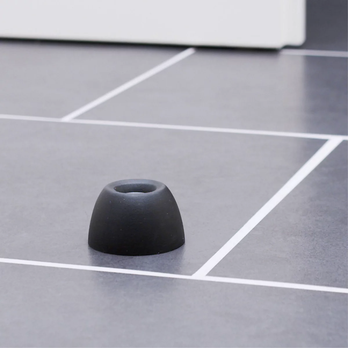 Un embout d'oreille noir repose sur une surface carrelée grise. Des lignes blanches forment un motif de grille.