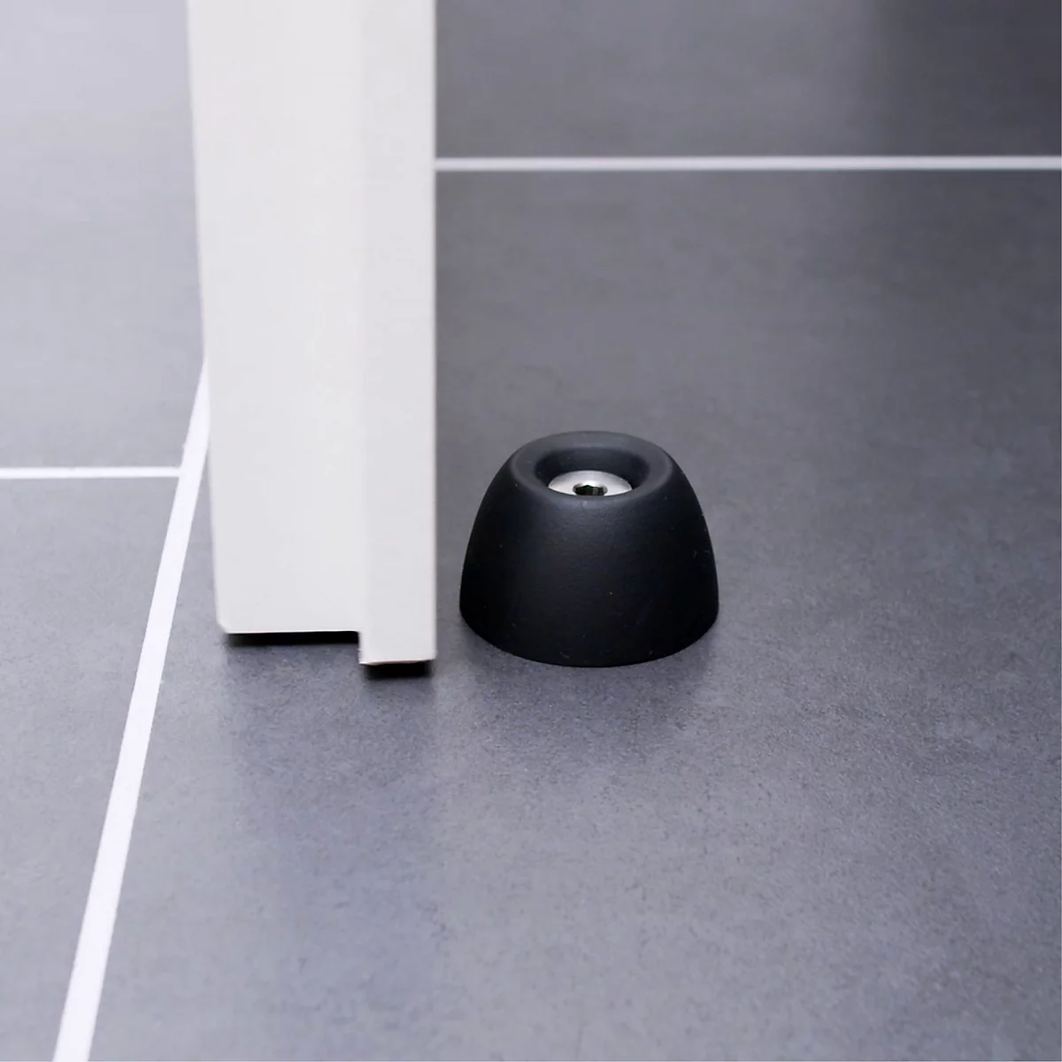 Un butoir de porte noir devant un cadre de porte blanc. Sur un sol gris. Le détail montre la forme de l'arrêt.