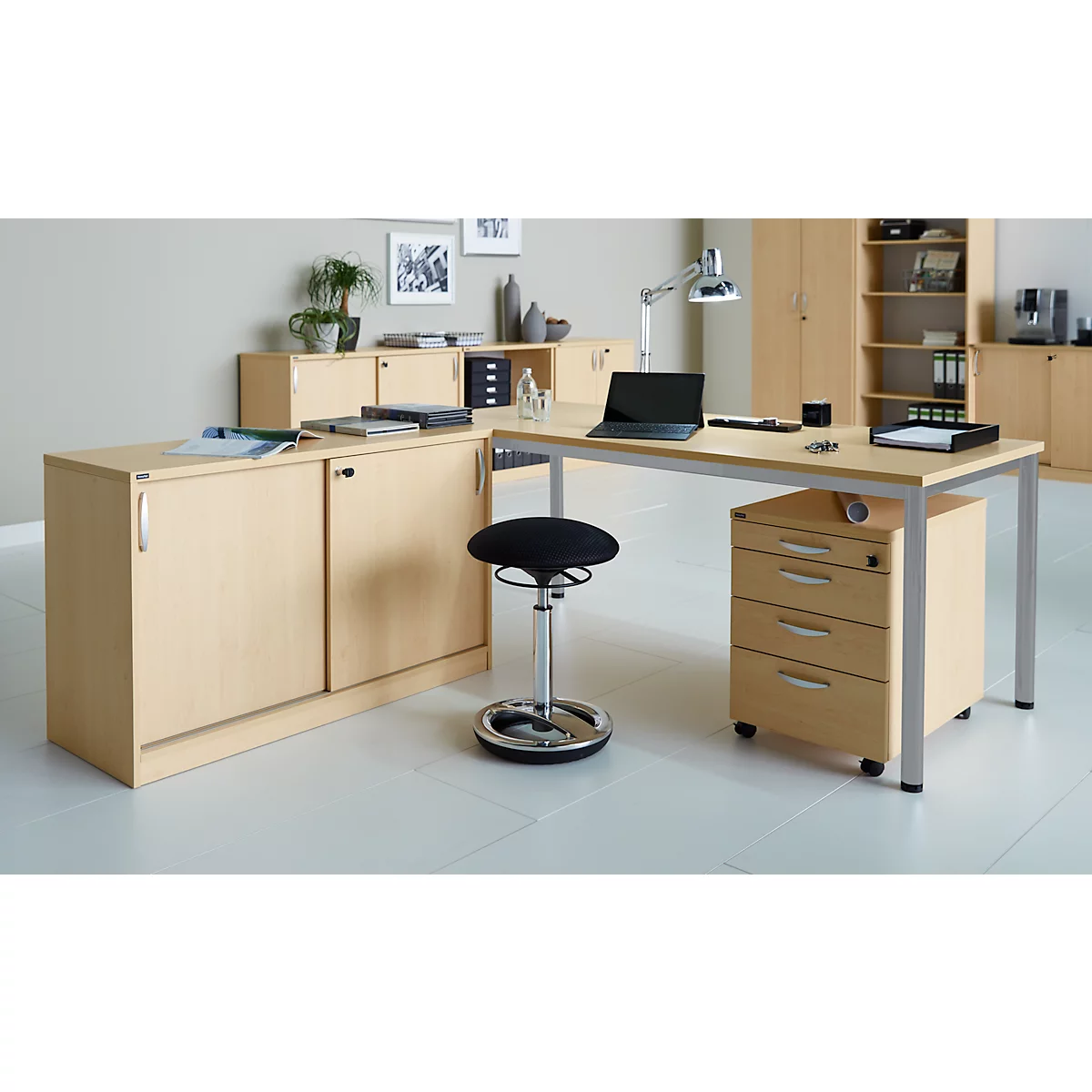 A bright office with a desk, mobile pedestal, stool, shelves, and plants.