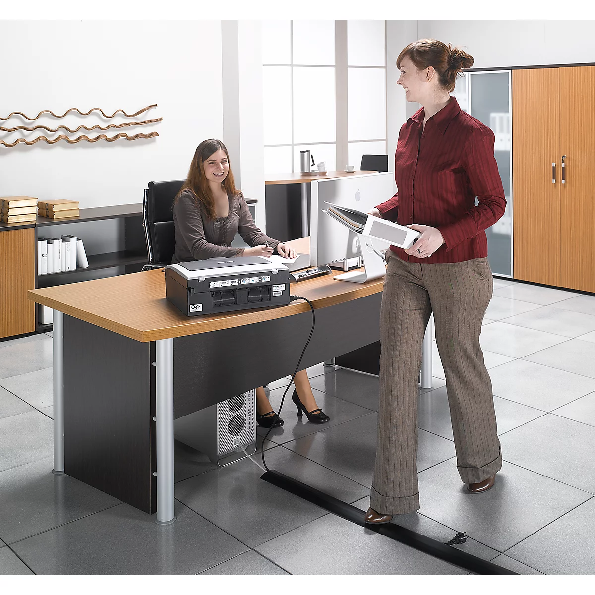 Two women in an office. One is sitting at the desk, the other is standing holding files. Office equipment.