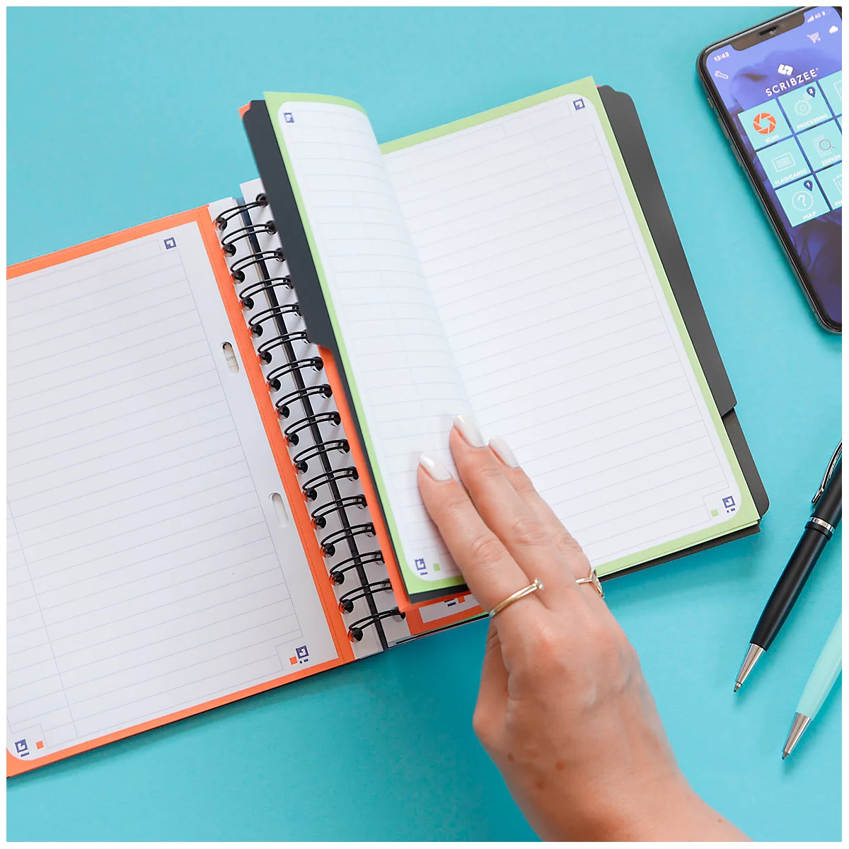 Gros plan : une personne feuillette un cahier avec des accents verts et oranges. Un téléphone et des stylos sont sur la table.