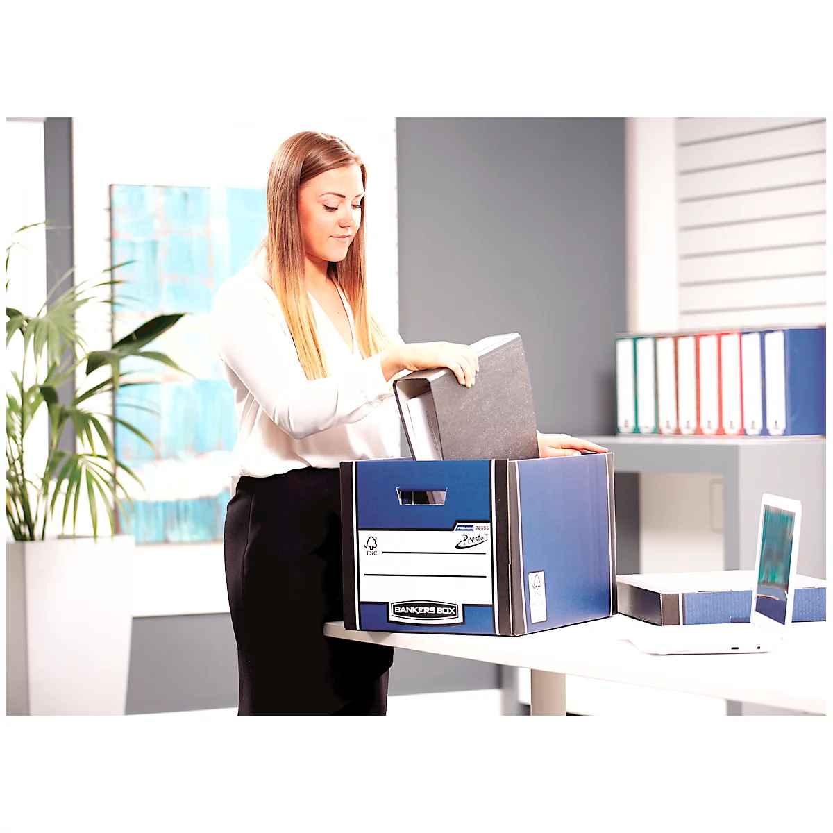 Une femme range des dossiers dans une boîte bleue. D'autres dossiers et dossiers sont visibles sur la table.