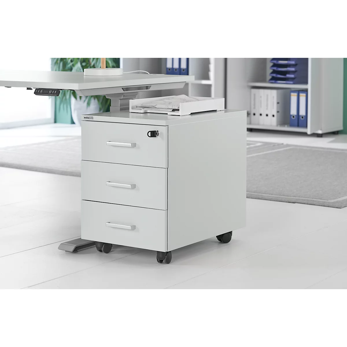 Caisson roulant gris avec trois tiroirs sous un bureau réglable en hauteur. Il y a un plateau sur le caisson.