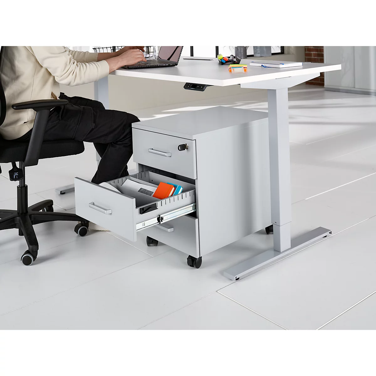 Une personne est assise à un bureau réglable en hauteur et travaille sur un ordinateur portable. Le tiroir du caisson mobile est ouvert.