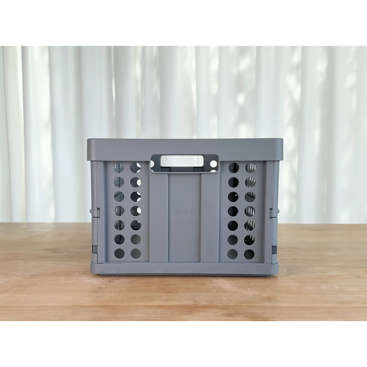 Caja plegable gris con agujeros, colocada sobre una mesa de madera frente a una cortina blanca.