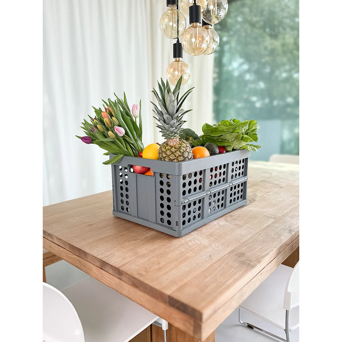 Caja gris llena de frutas y flores sobre una mesa de madera, con una lámpara colgando encima. Dos sillas están preparadas.