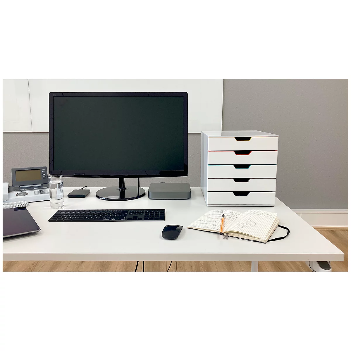 Un escritorio con una computadora, un gabinete de cajones, un cuaderno y un ratón. Monitor gris, gabinete blanco.