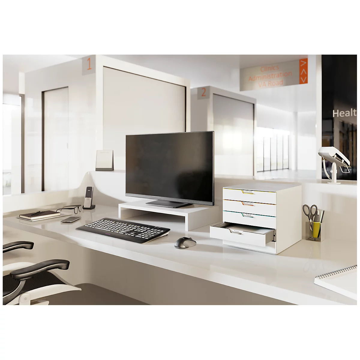 Un mostrador de recepción moderno con ordenador, mueble cajonero, teléfono y portalápices. En el fondo se ven ventanas y señalización.