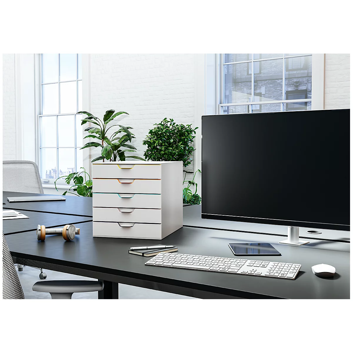 Escritorio de oficina con sistema de cajones blanco, monitor, teclado, bolígrafos y plantas.