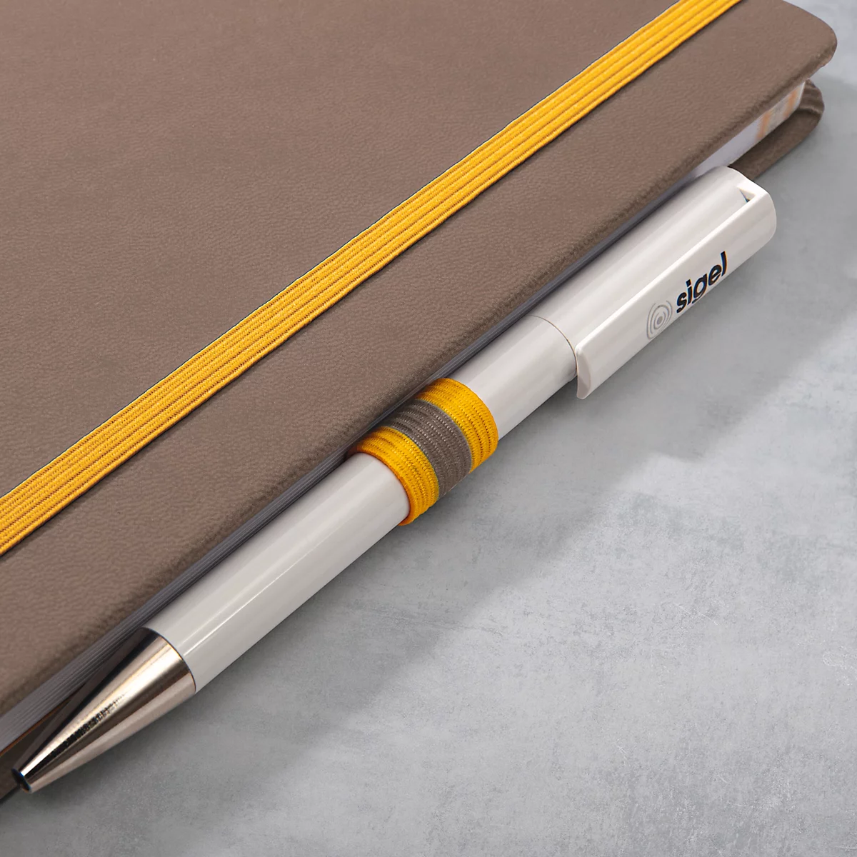 Cahier gris avec des bandes élastiques jaunes. Stylo blanc avec impression "sigel" dans le livre.