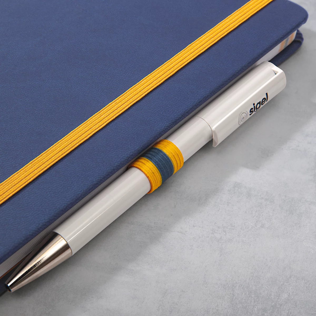 Stylo inséré dans un cahier bleu. Le stylo est blanc avec une bande bleue et jaune, le cahier a une bande élastique jaune.