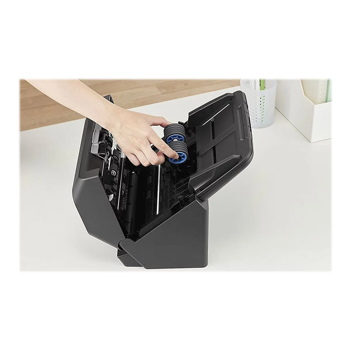 Eine Hand hält eine blaue Rolle in einem geöffneten schwarzen Scanner. Auf hellem Tisch, Behältnisse im Hintergrund.
