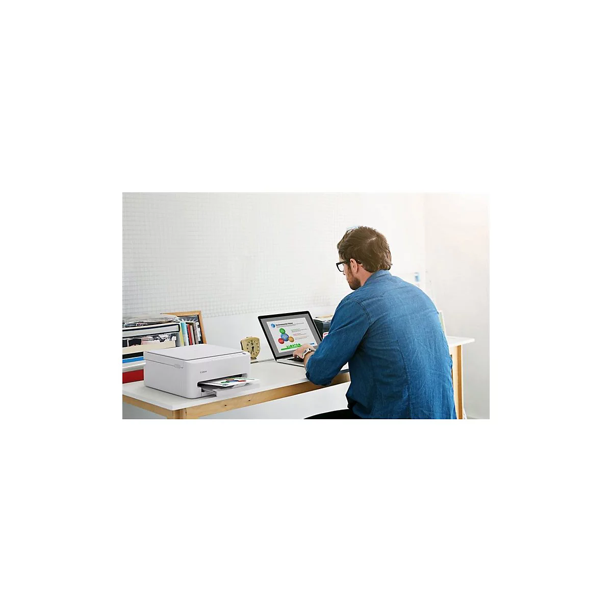Ein Mann sitzt an einem Schreibtisch und arbeitet mit einem Laptop und einem Drucker. Der Drucker druckt Papier aus.