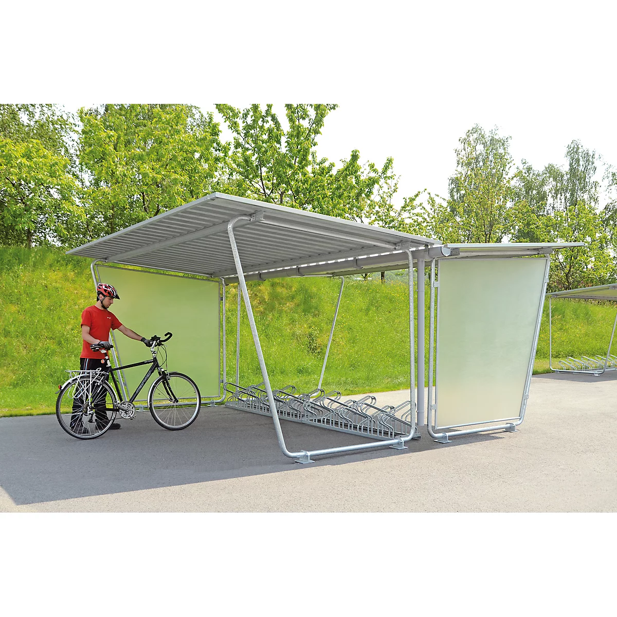 Person with a bicycle under a metal bike shelter. A green wall is behind the person. Trees in the background.