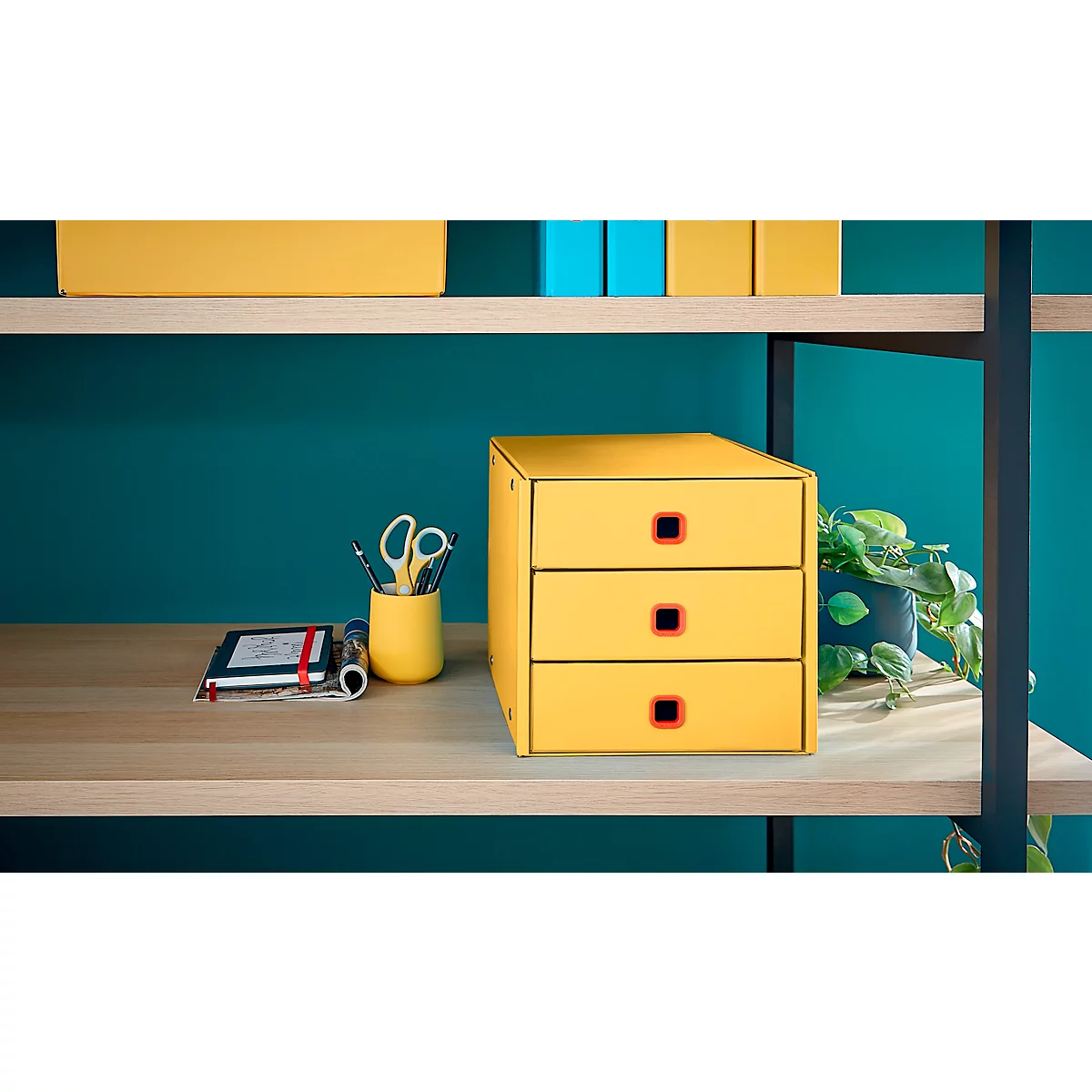 Boîte à tiroirs jaune avec bouton rouge, ciseaux, porte-stylo, carnet et plante sur une étagère en bois.