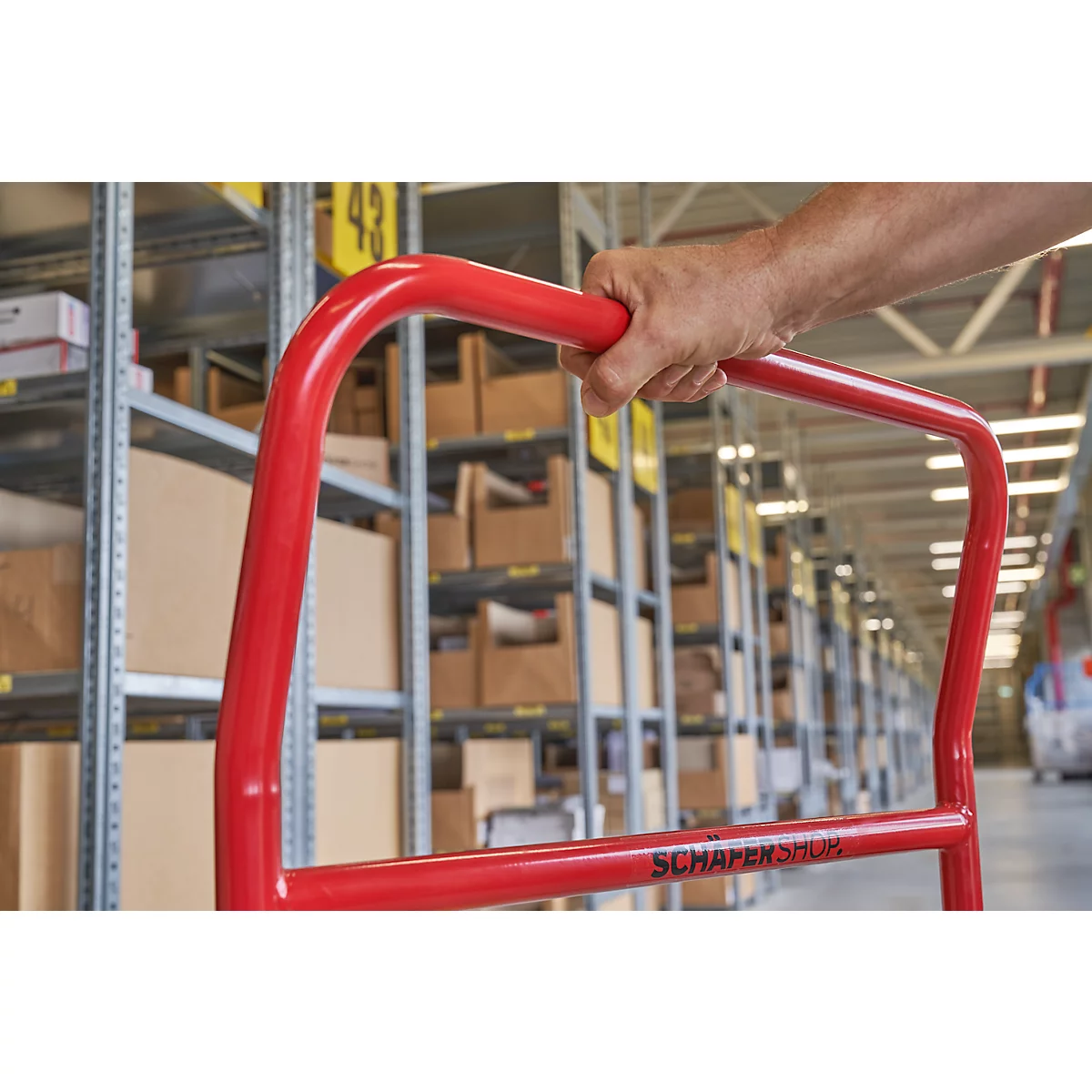 Carretilla roja en un almacén. Un brazo sostiene el asa. Estanterías con cajas al fondo.