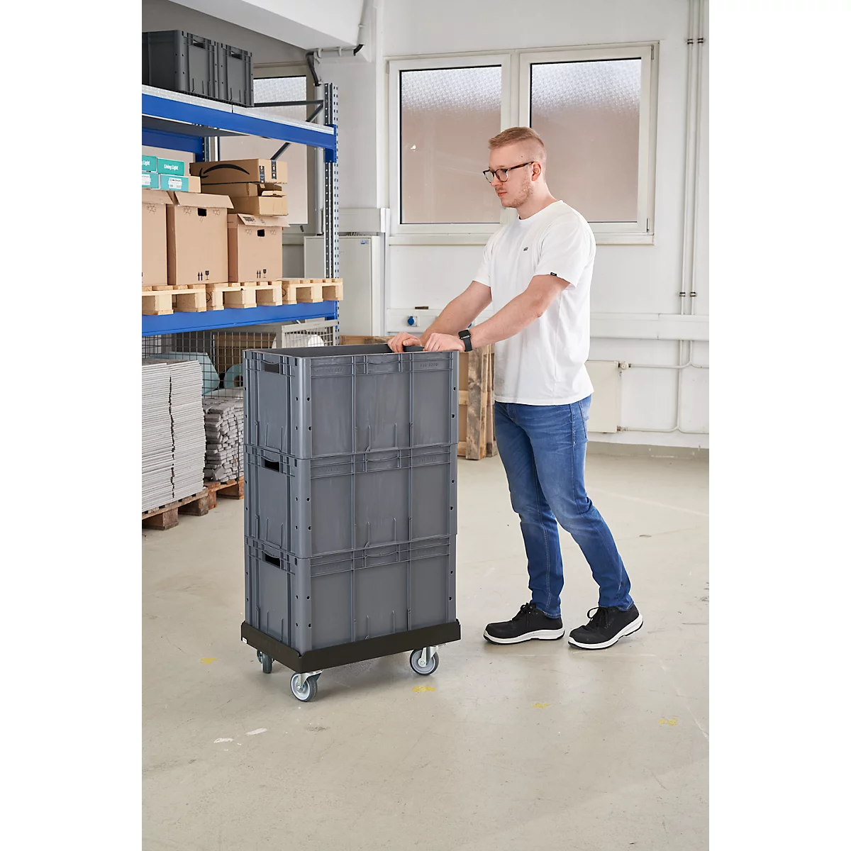 Un hombre empuja un contenedor de transporte gris con ruedas. En el fondo, estanterías con cajas.