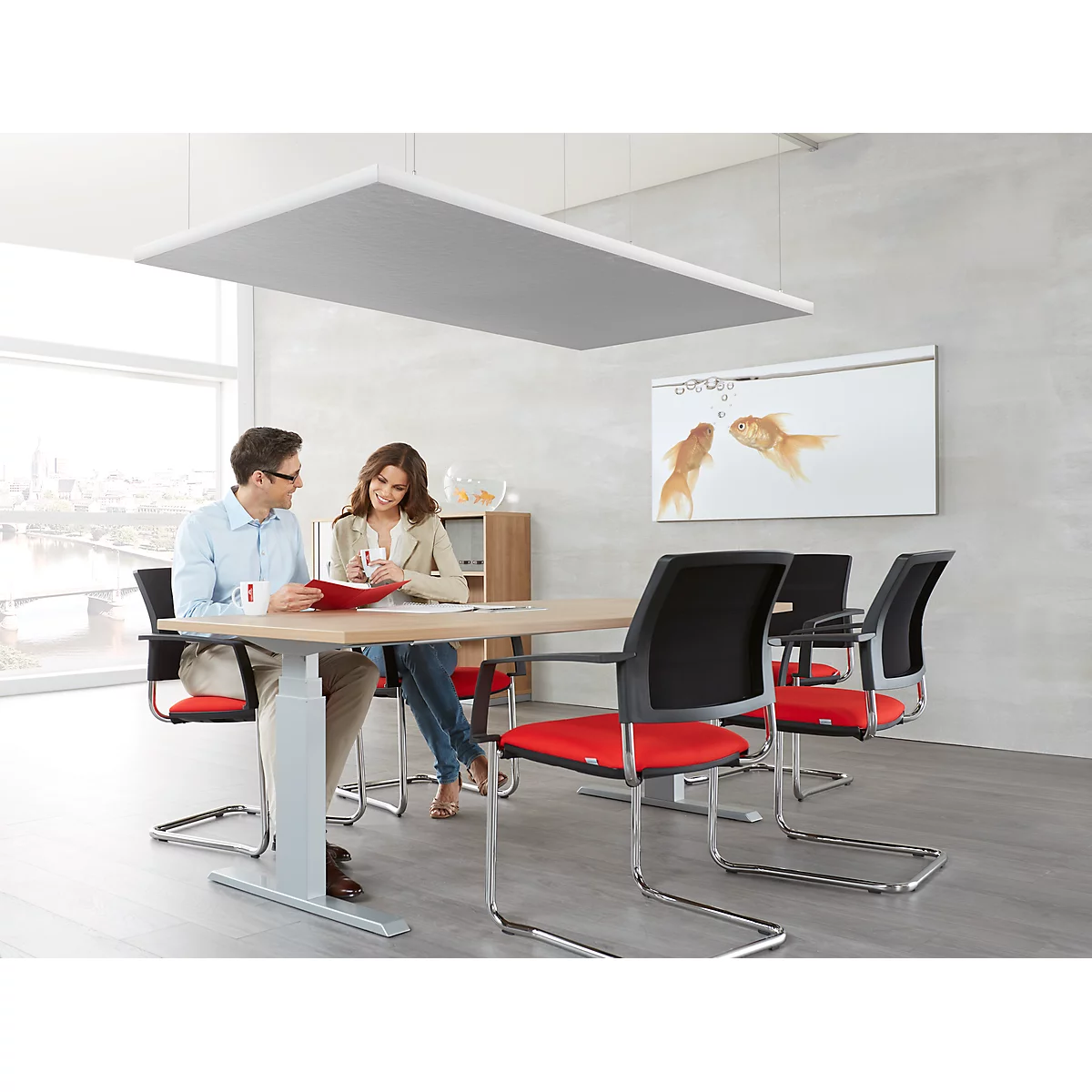 Two people sit at a table in a bright conference room. A work of art with two goldfish hangs on the wall.
