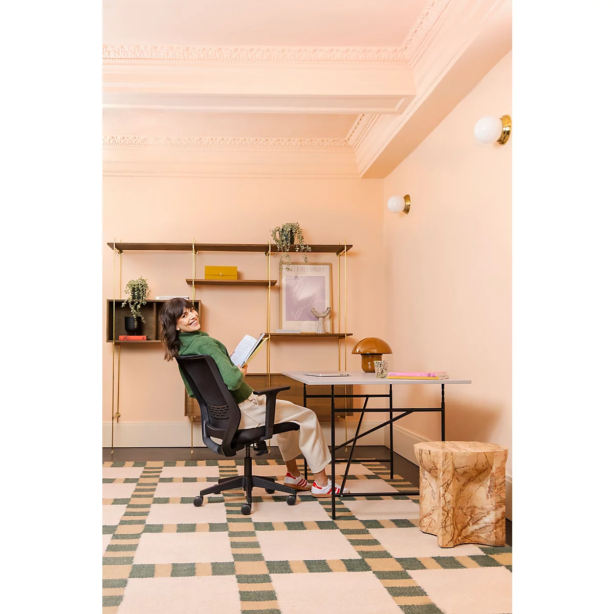 Une femme souriante se penche en arrière sur une chaise de bureau, lisant un livre. La pièce est lumineuse et attrayante.