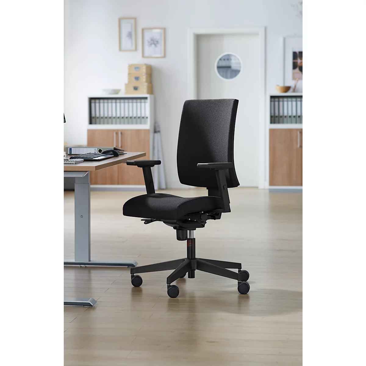 Un fauteuil de bureau noir dans un bureau lumineux. La chaise a un dossier haut et des accoudoirs.