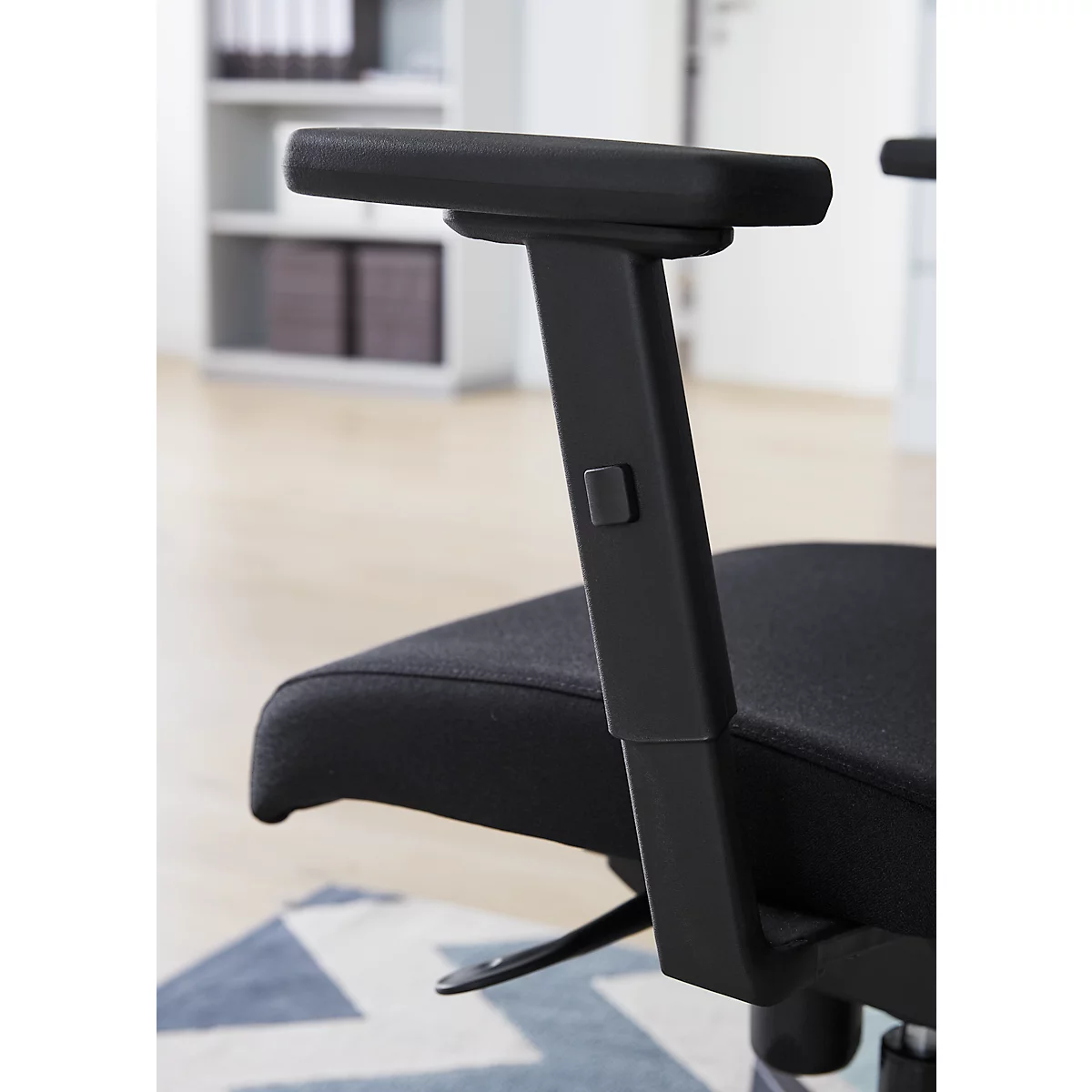 Gros plan sur une chaise de bureau noire. Accoudoir, assise et partie du dossier. Arrière-plan : étagère et tapis.