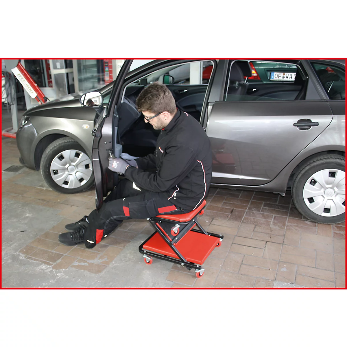 Un homme en vêtements de travail répare une voiture, assis sur un tabouret roulant rouge.