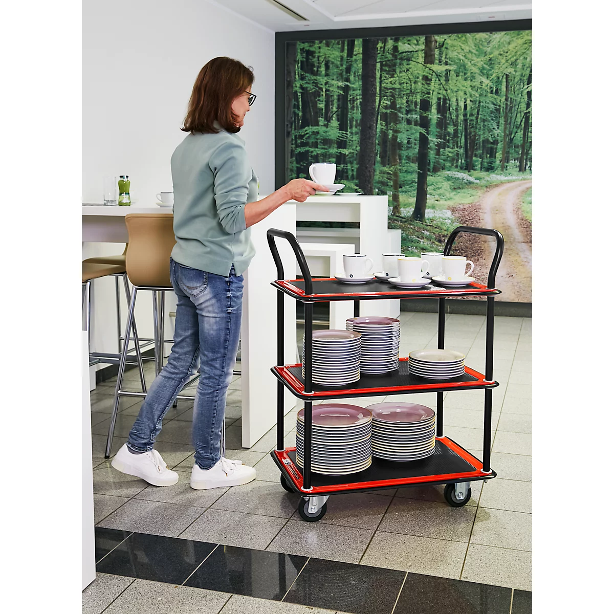 Femme portant des lunettes, place une tasse sur un chariot de service avec de la vaisselle. Paysage forestier en arrière-plan.