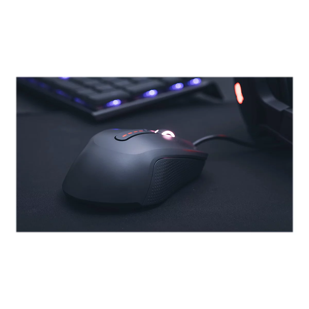 Schwarze Gaming-Maus auf schwarzem Untergrund. Im Hintergrund eine Tastatur und ein Headset.