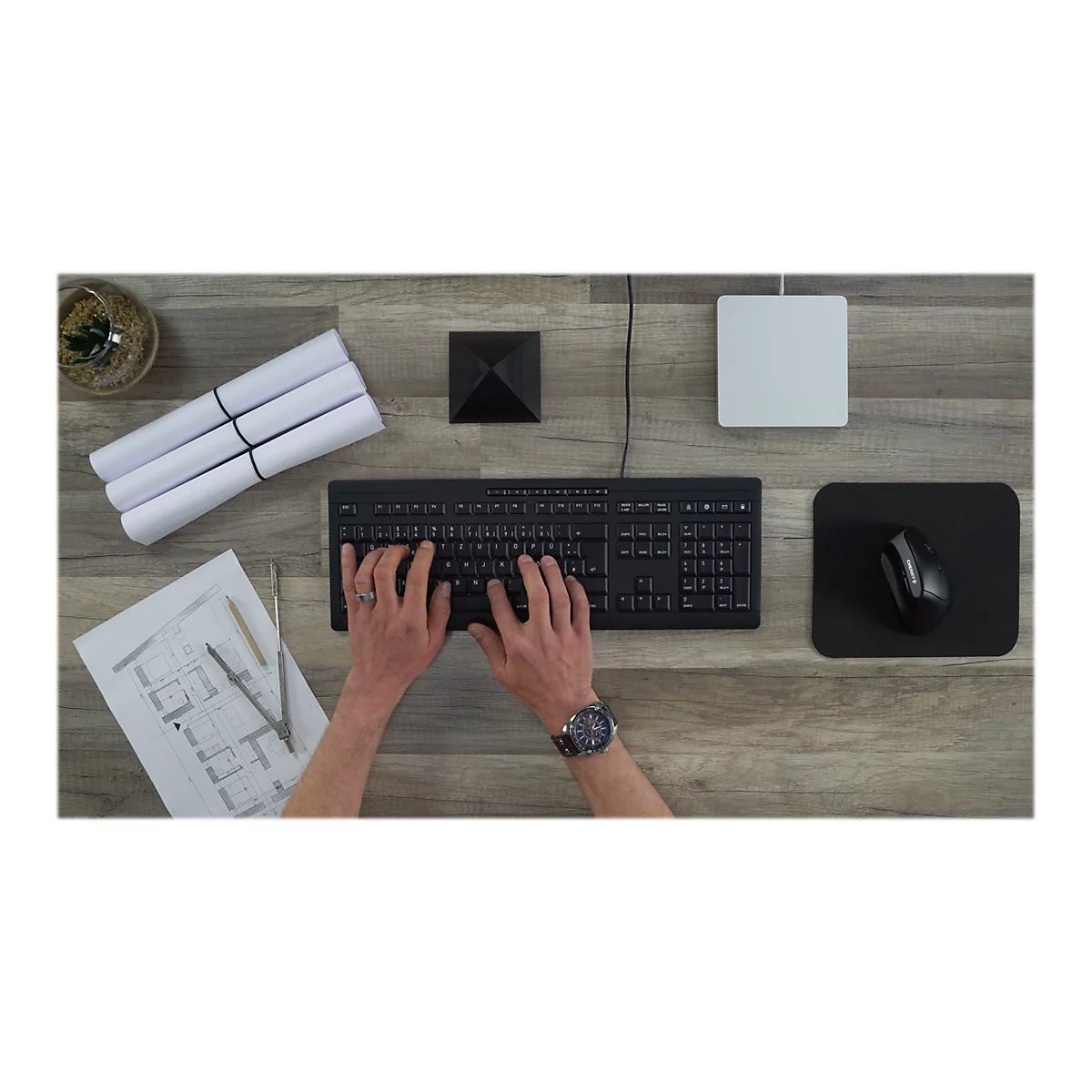 Person tippt auf schwarzer Tastatur. Daneben liegen ein Mauspad, eine Maus, PlÃ¤ne und ein weiÃŸes KÃ¤stchen.