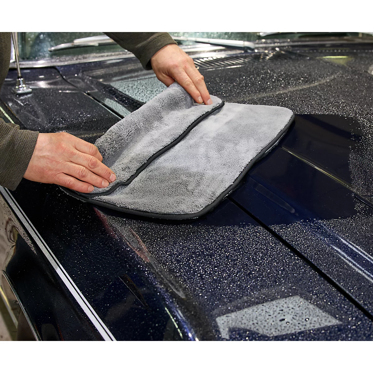 Les mains essuient les gouttelettes d'eau du capot bleu d'une voiture avec un chiffon en microfibre gris.