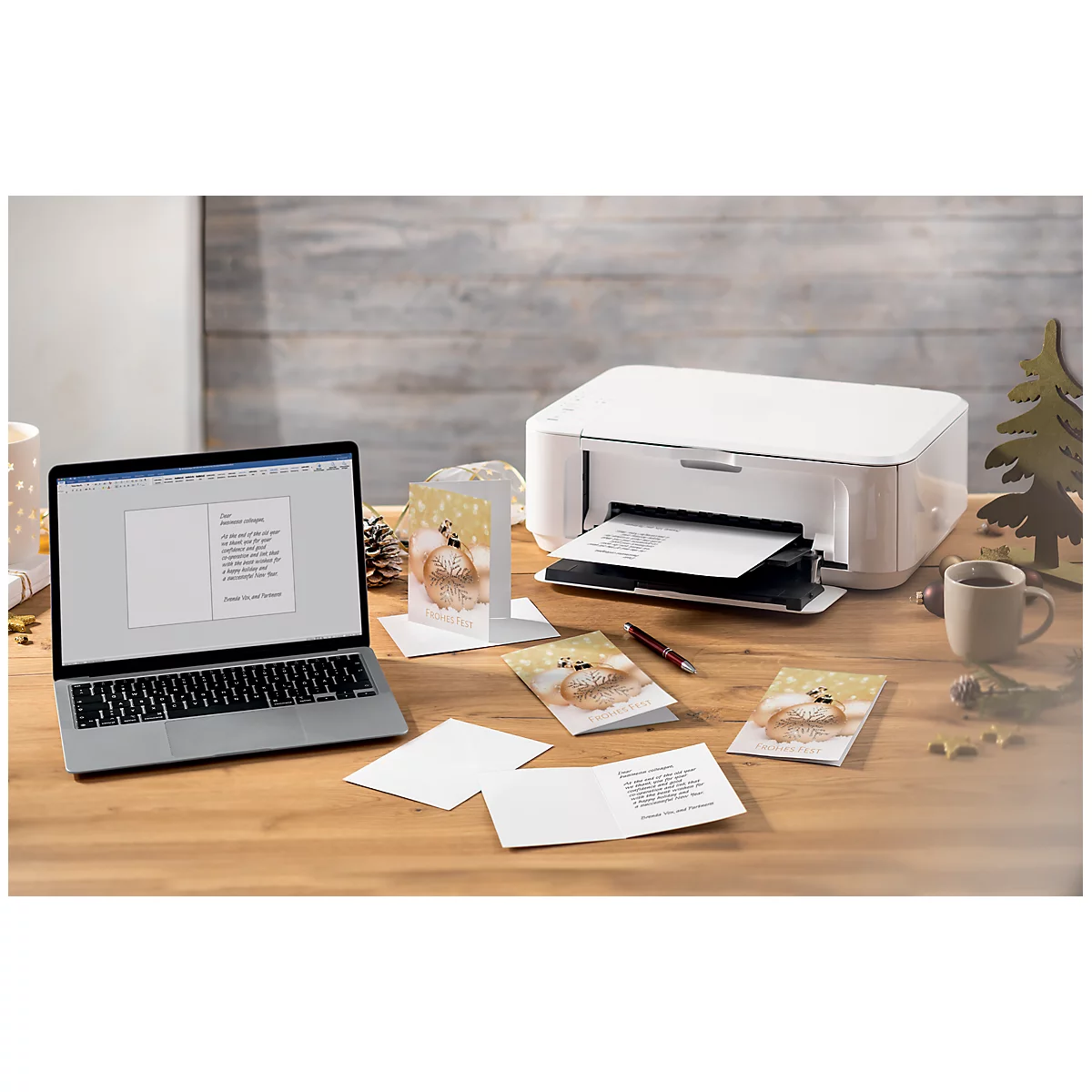 Laptop, printer, and Christmas cards on a wooden table. The screen displays text being printed. Several finished cards are beside it.