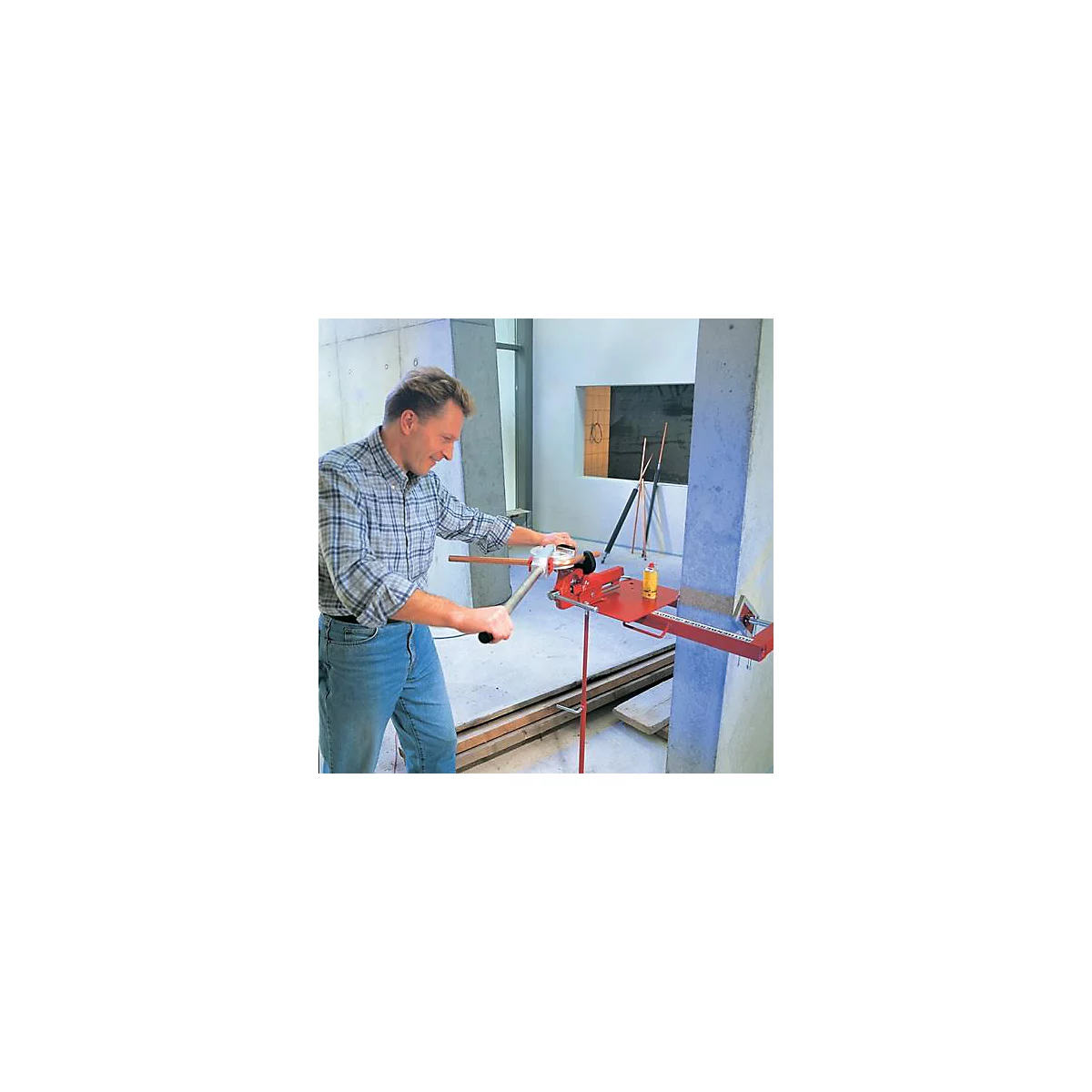 Un homme plie un tuyau en cuivre à l'aide d'un outil rouge. Il porte un jean et une chemise à carreaux dans un atelier lumineux.