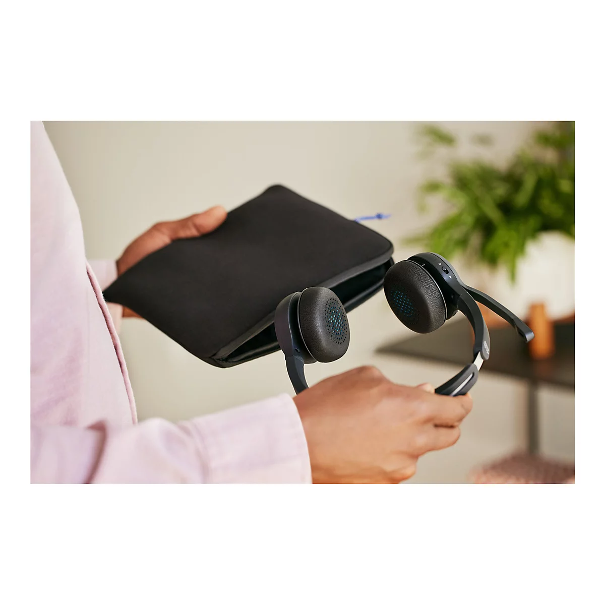 Person hält schwarzes Headset und eine Tasche. Das Headset hat gepolsterte Ohrhörer und einen schwarzen Bügel.