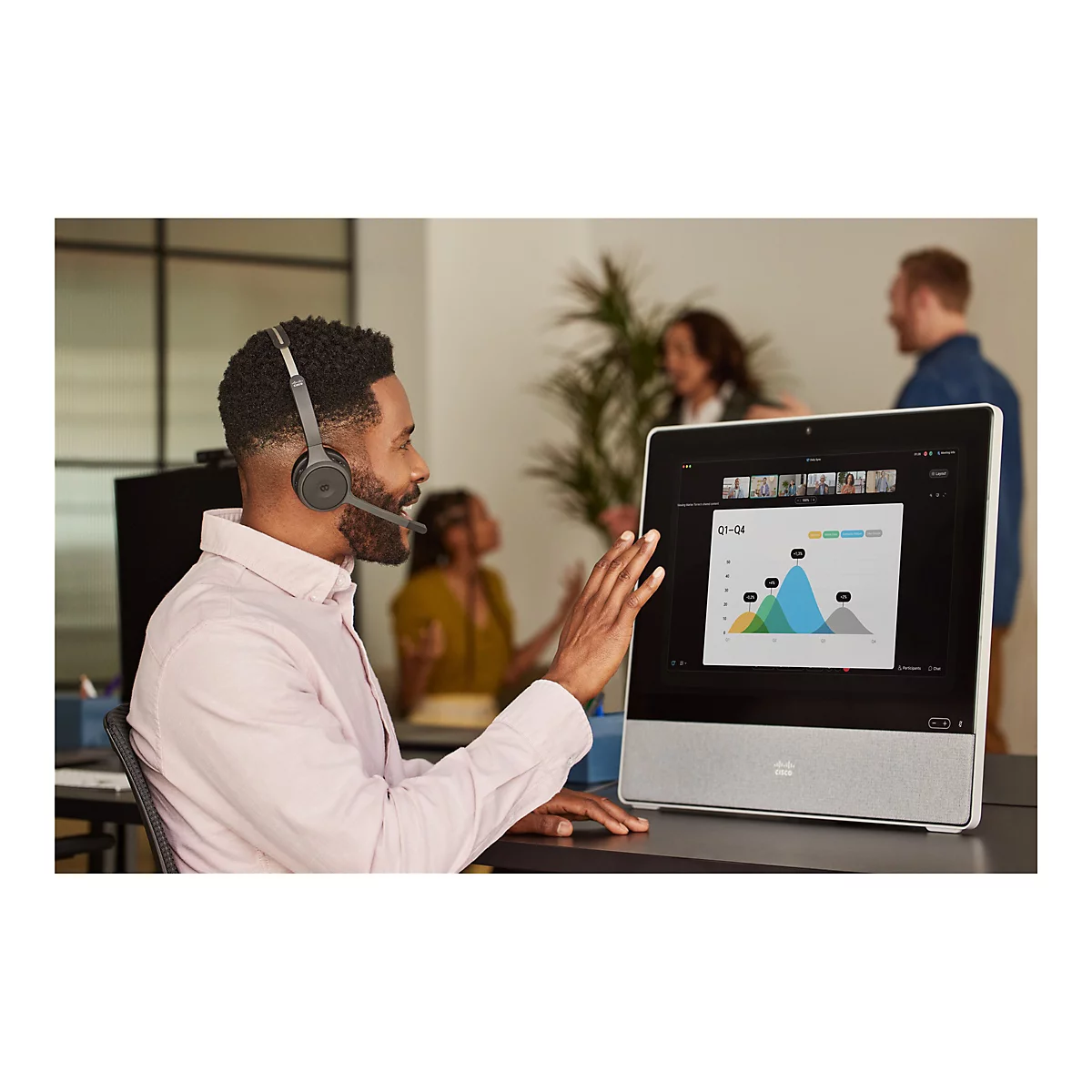 Ein Mann mit Headset interagiert per Touchscreen mit einem Videokonferenz-Gerät. Im Hintergrund sind Kollegen zu sehen.