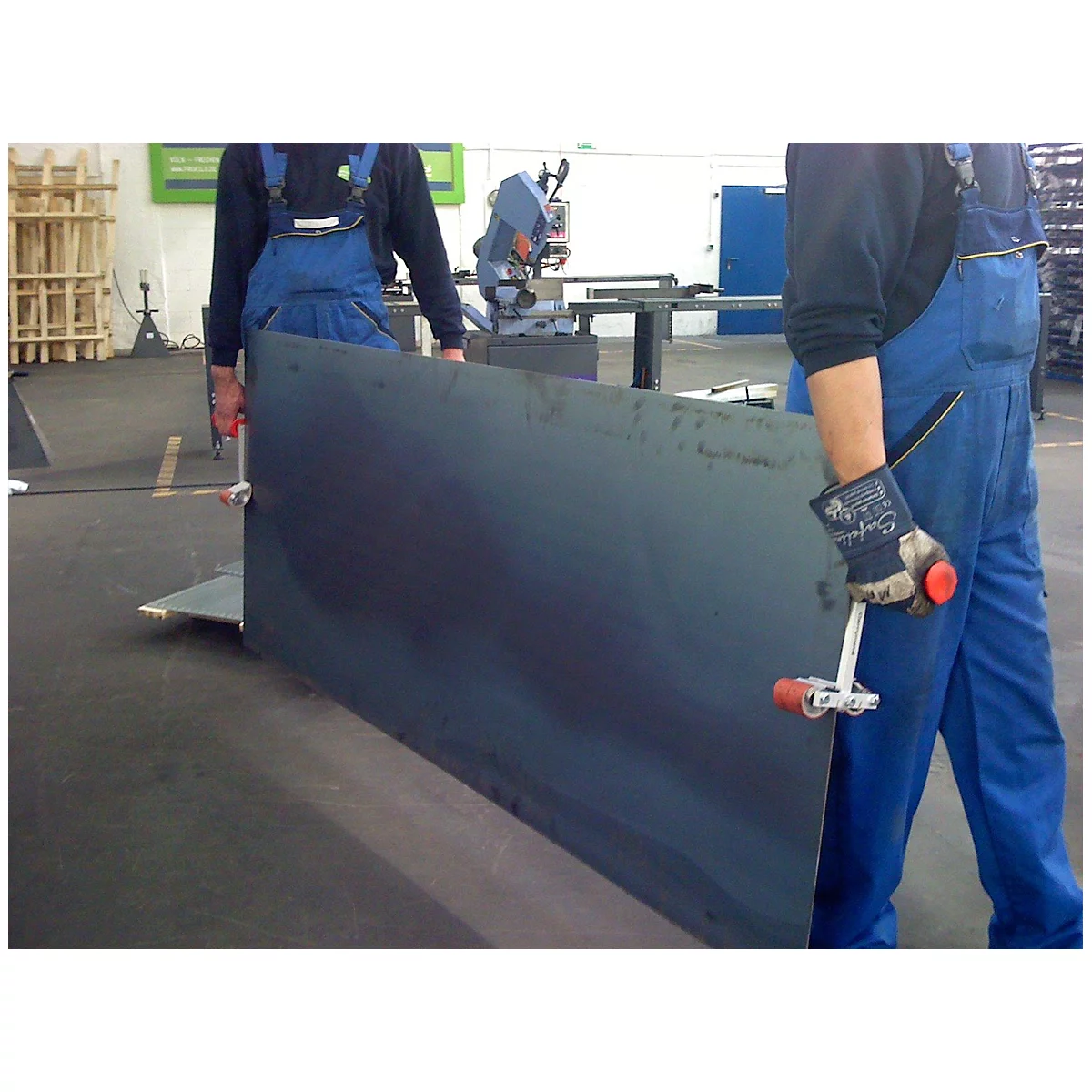Two workers carrying a large steel plate in a factory. One has a white gripper with a red handle in his hand.