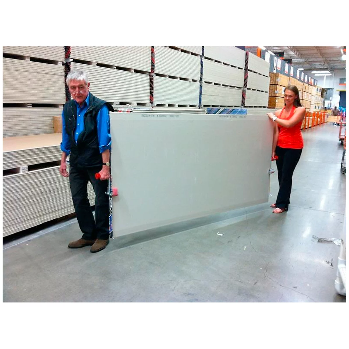 A man and woman carrying a large drywall sheet together in a hardware store.