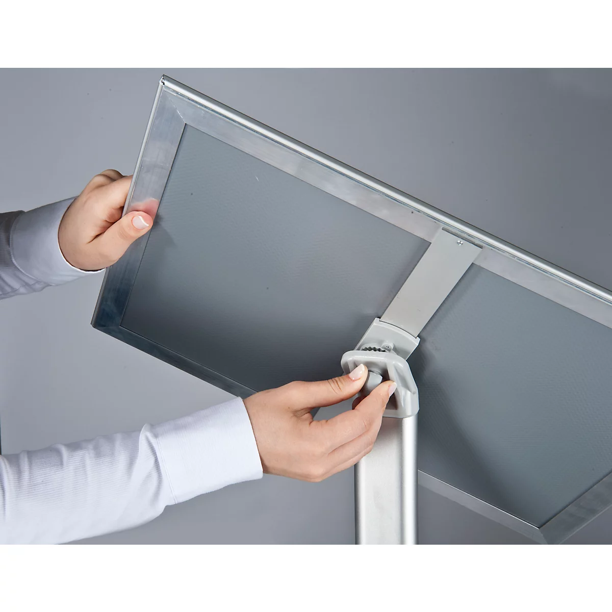 Hands assembling a grey display, attaching it to a metal frame. Close-up on a grey background.