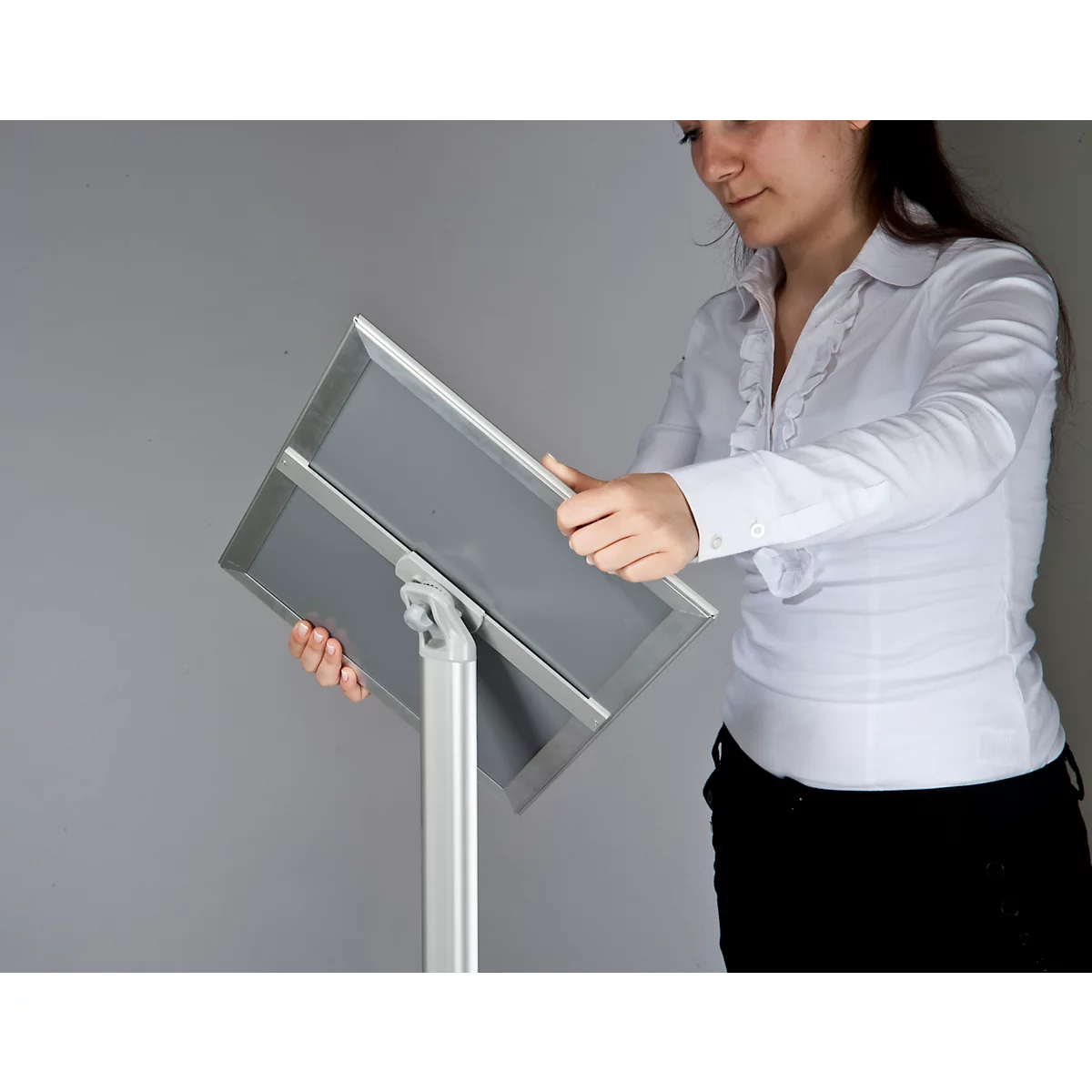 A woman holds an information stand. She is wearing a white shirt and black trousers. The stand is made of metal.
