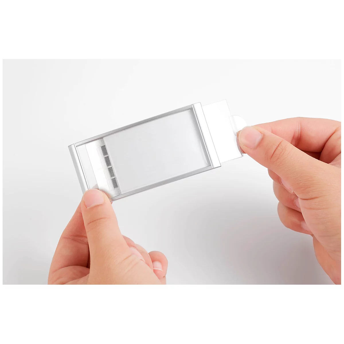 Hands holding a light gray rectangular object with transparent window. The object frame is silver, hands against a white background.