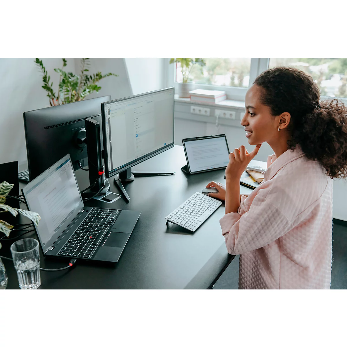 Une femme est assise à un bureau, travaillant sur un ordinateur, une tablette, un clavier et souriant.