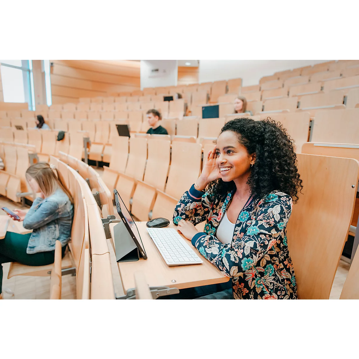 Une femme souriante est assise dans un amphithéâtre. Elle a les cheveux bouclés et porte une veste à motifs floraux. Il y a une tablette, un clavier et une souris sur le bureau.
