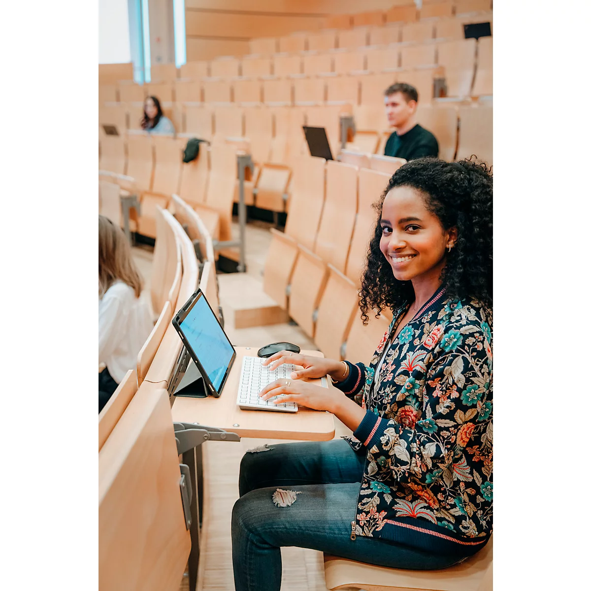 Une jeune femme aux cheveux bouclés sourit dans un amphithéâtre tout en travaillant sur un ordinateur portable. D'autres étudiants sont en arrière-plan.
