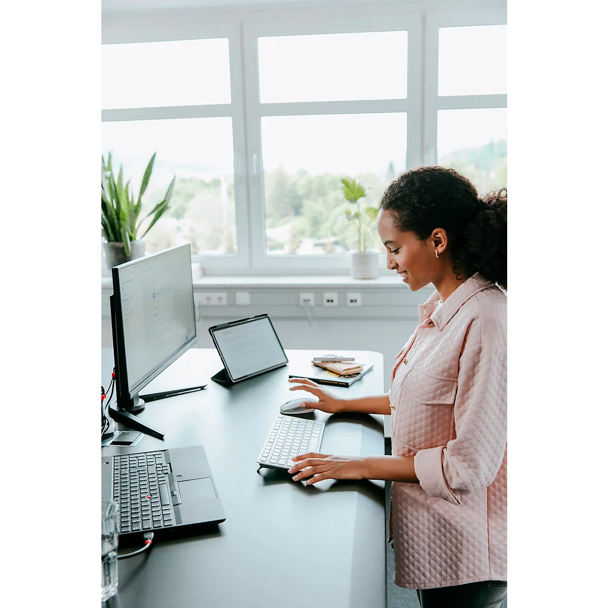 Une femme est assise à un bureau. Elle tape sur un clavier, à côté d'un moniteur et d'une tablette. Derrière elle, il y a une fenêtre avec des arbres.