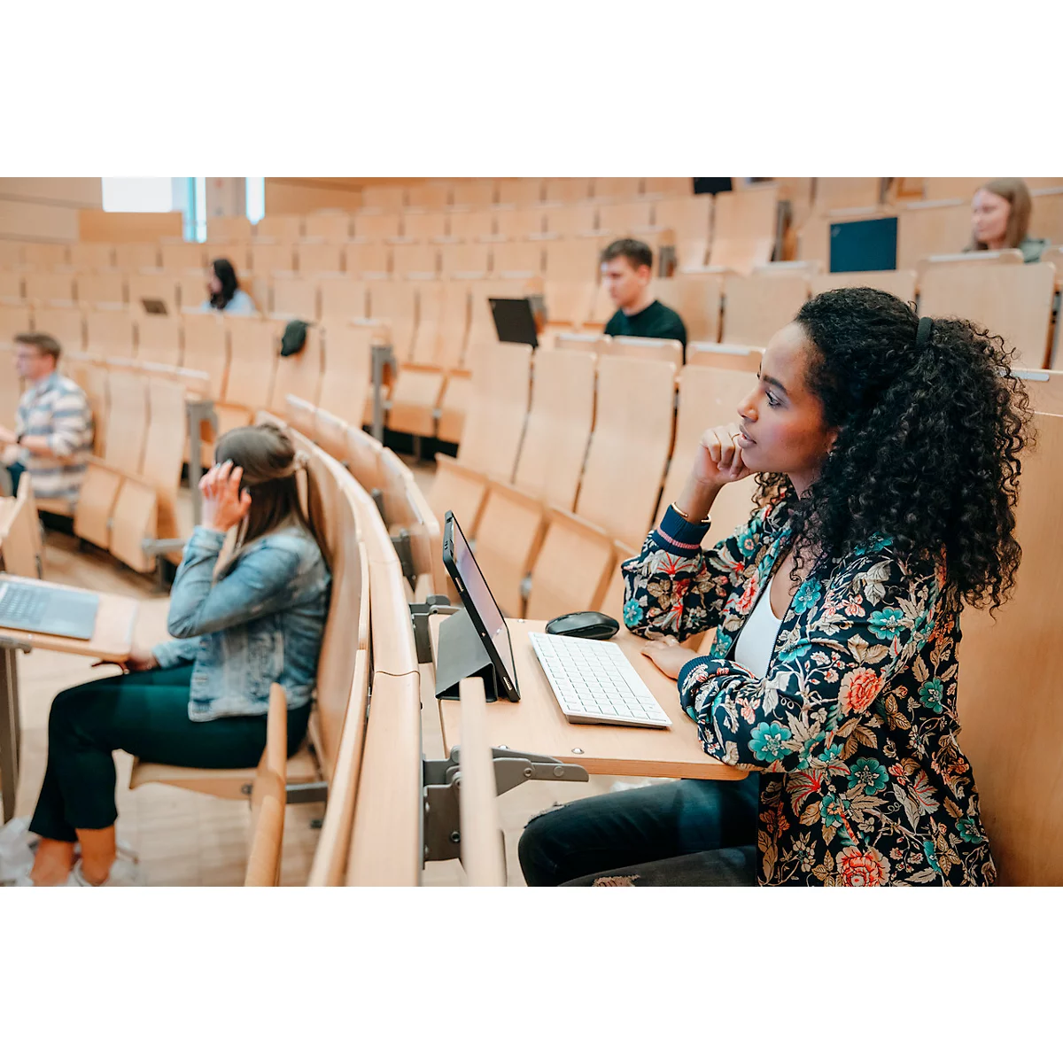 Une étudiante aux cheveux bouclés dans un amphithéâtre, à un pupitre avec une tablette. D'autres étudiants sont visibles en arrière-plan.