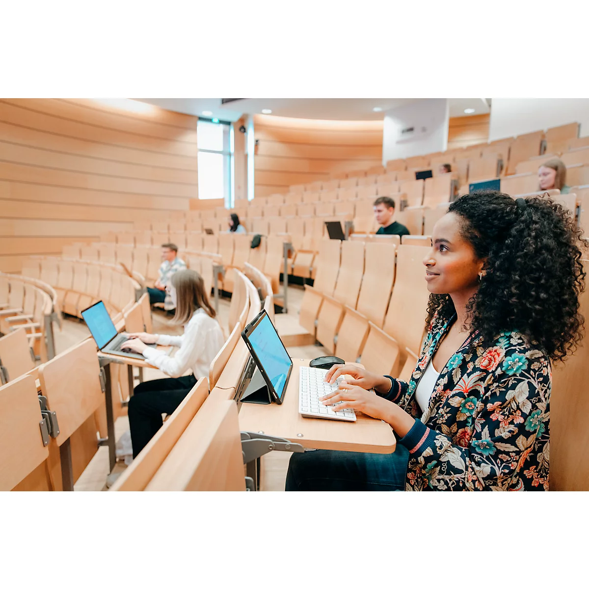 Jeune femme aux cheveux bouclés dans un amphithéâtre tapant sur un clavier. D'autres étudiants sont en arrière-plan.