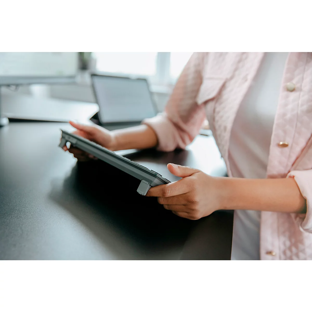 Une femme tient une tablette grise dans ses mains. Elle porte une veste rose. En arrière-plan, on peut voir un autre ordinateur portable et une table.